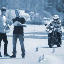 Motorcyclist takes the license exam while two men discuss results